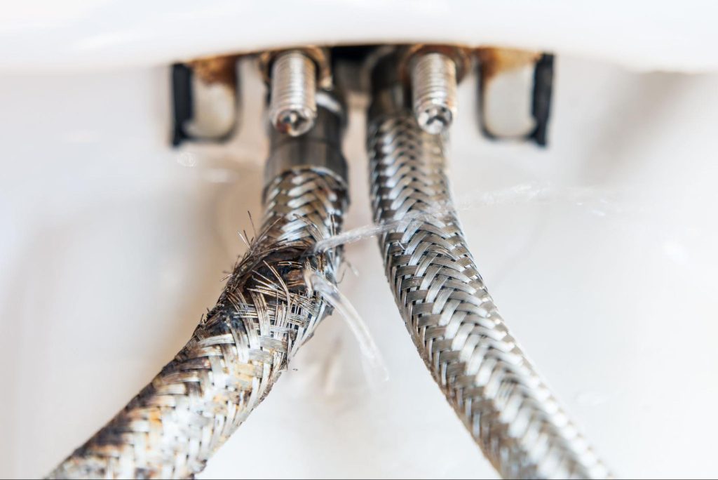 Damaged braided stainless steel water supply hose under a sink, with frayed outer mesh and visible leak spraying water